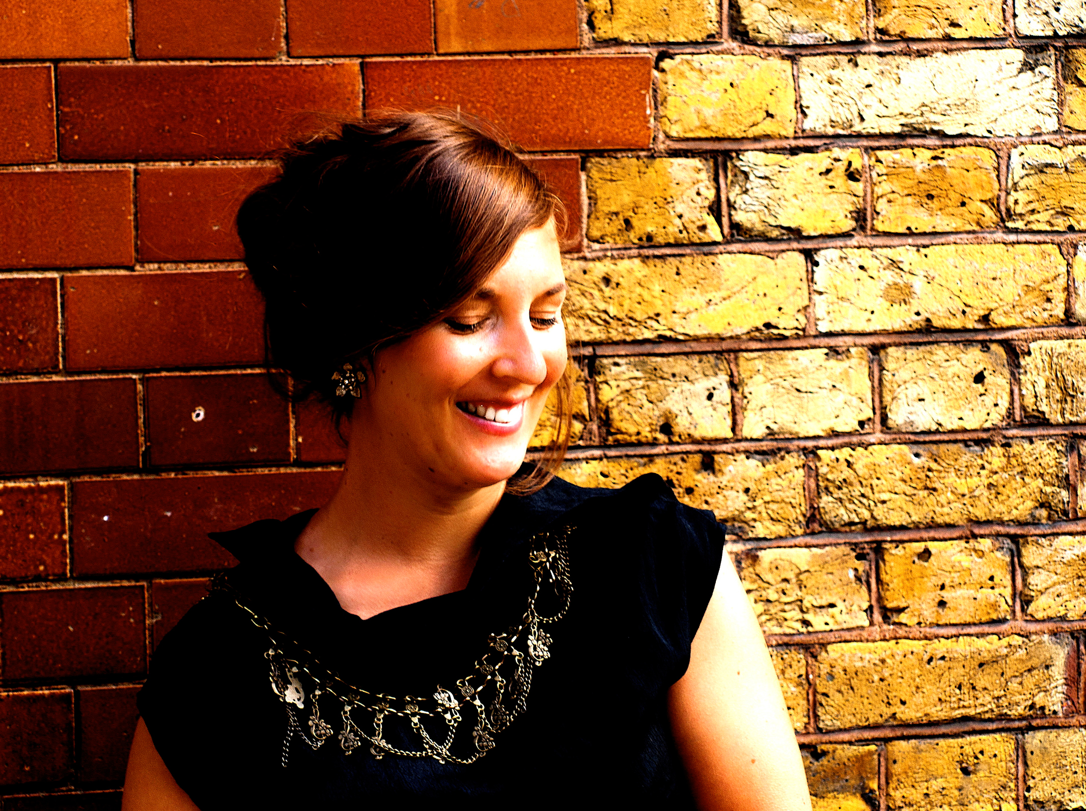Conductor Kelly Lovelady stands against a brick wall of two distinct textures in a flowing black top and oversized jewellery. On the left, the diagonal sweep of her hair continues into a smooth red-brown brickwork. On the right, the light on her face continues into a grittier yellow wall